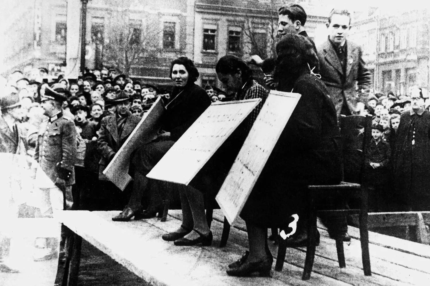 A crowd and uniformed Nazi officers look upon three Jewish women they’ve forced to sit on a platform and hold cardboard signs. 