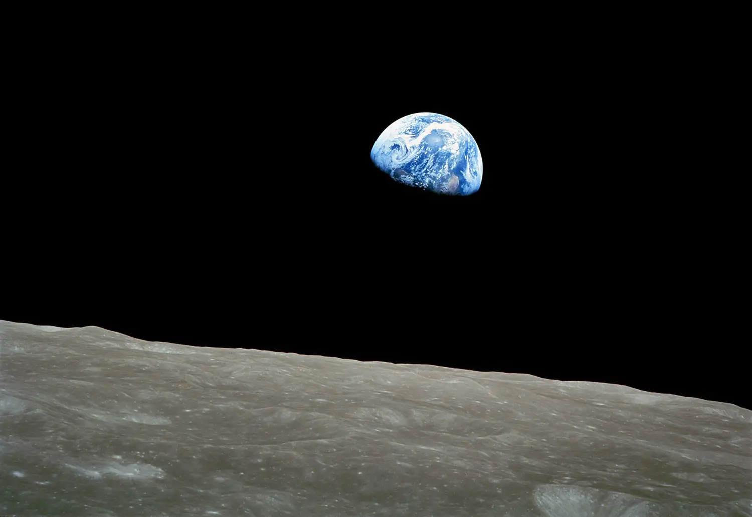 A crescent Earth is seen hovering, small, over the barren gray horizon of the moon.