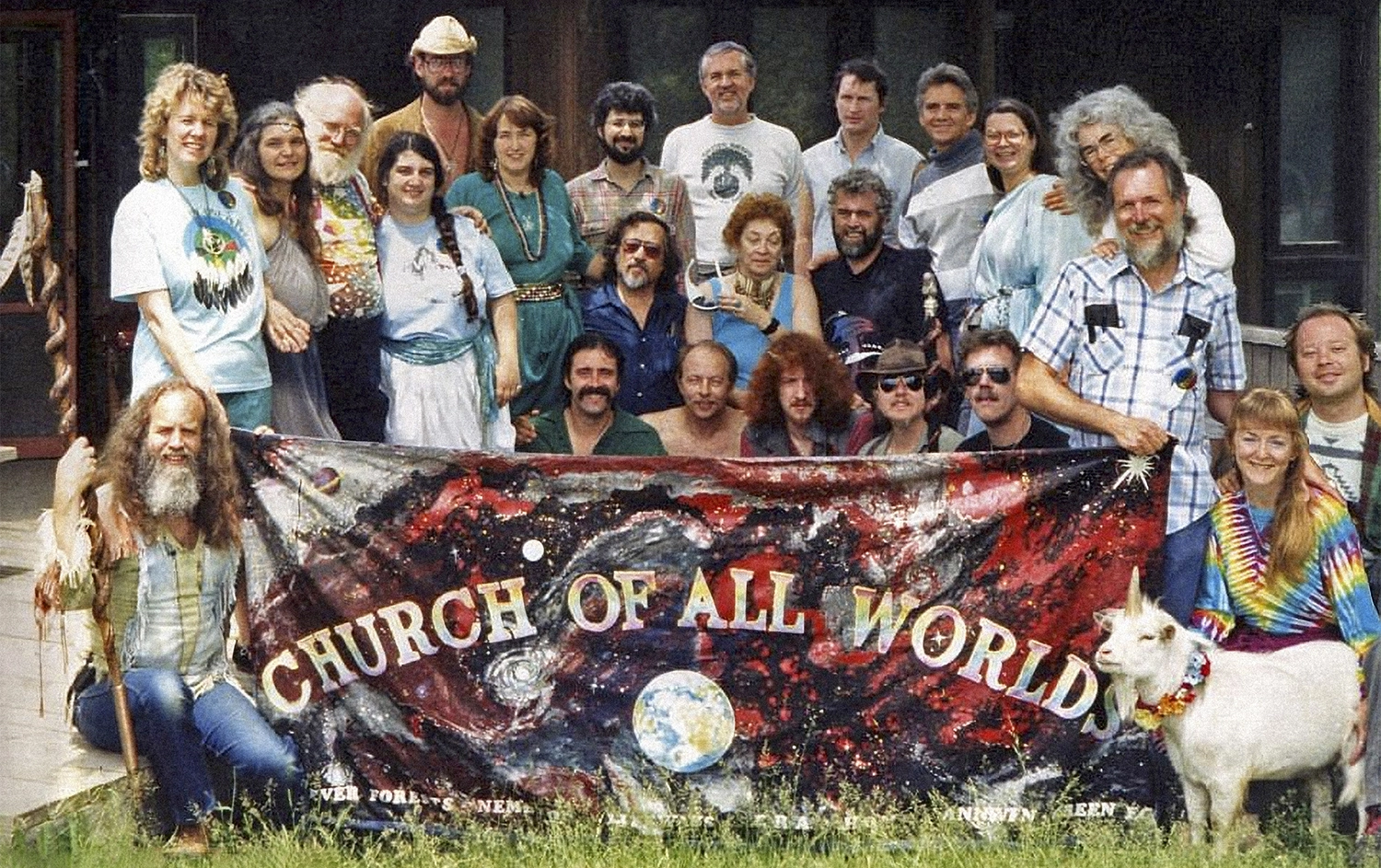 A group photo of the Church of All Worlds. They are holding a banner that says “Church of All Worlds” and there is also a white goat with a single horn, looking like a little unicorn.