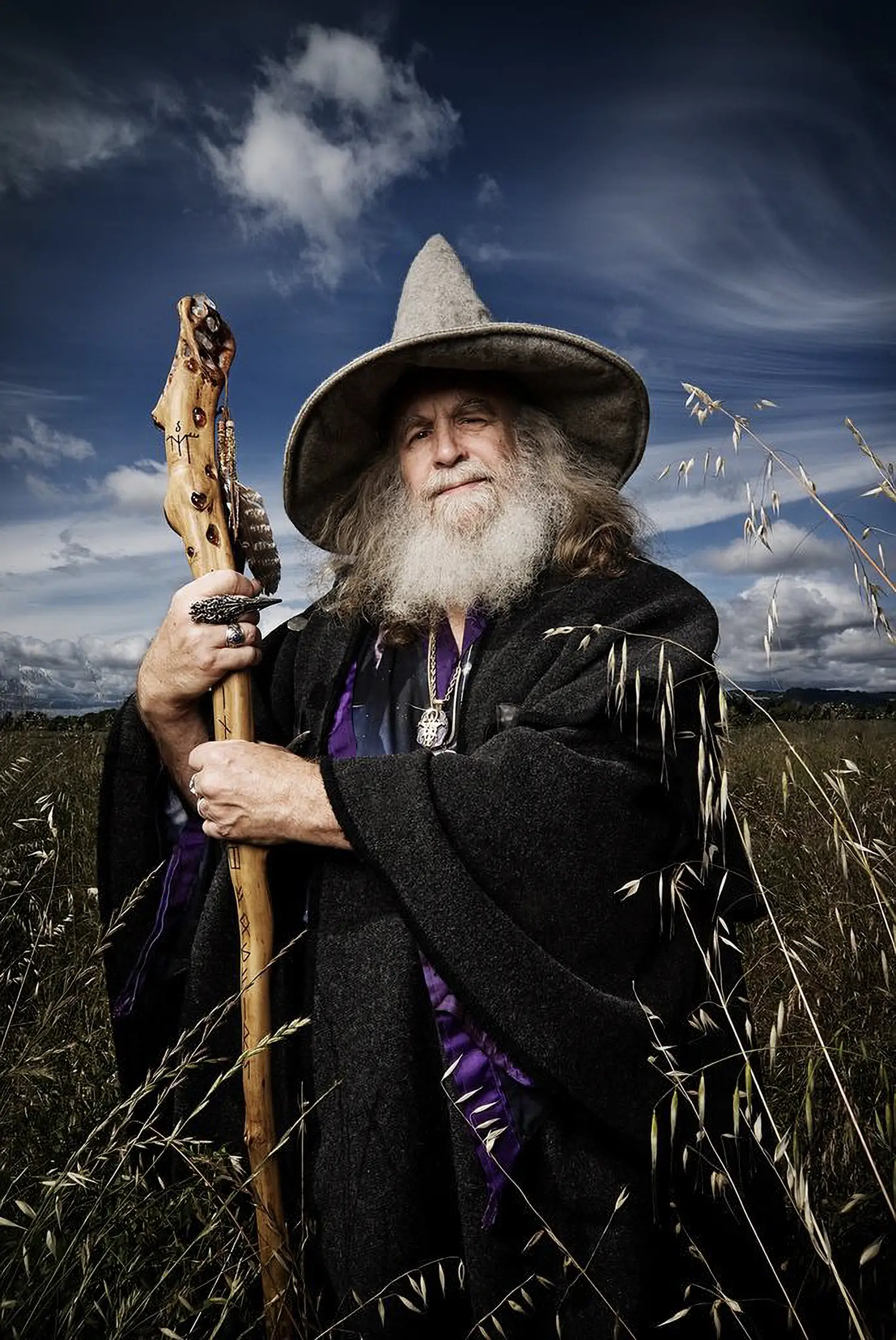 Portrait of Oberon Zell. He has flowing white hair, standing majestically in a field, holding a carved wooden staff. Dressed in a black cloak and wide-brimmed hat, his attire is accented with deep purple fabric and adorned with various trinkets and symbols.