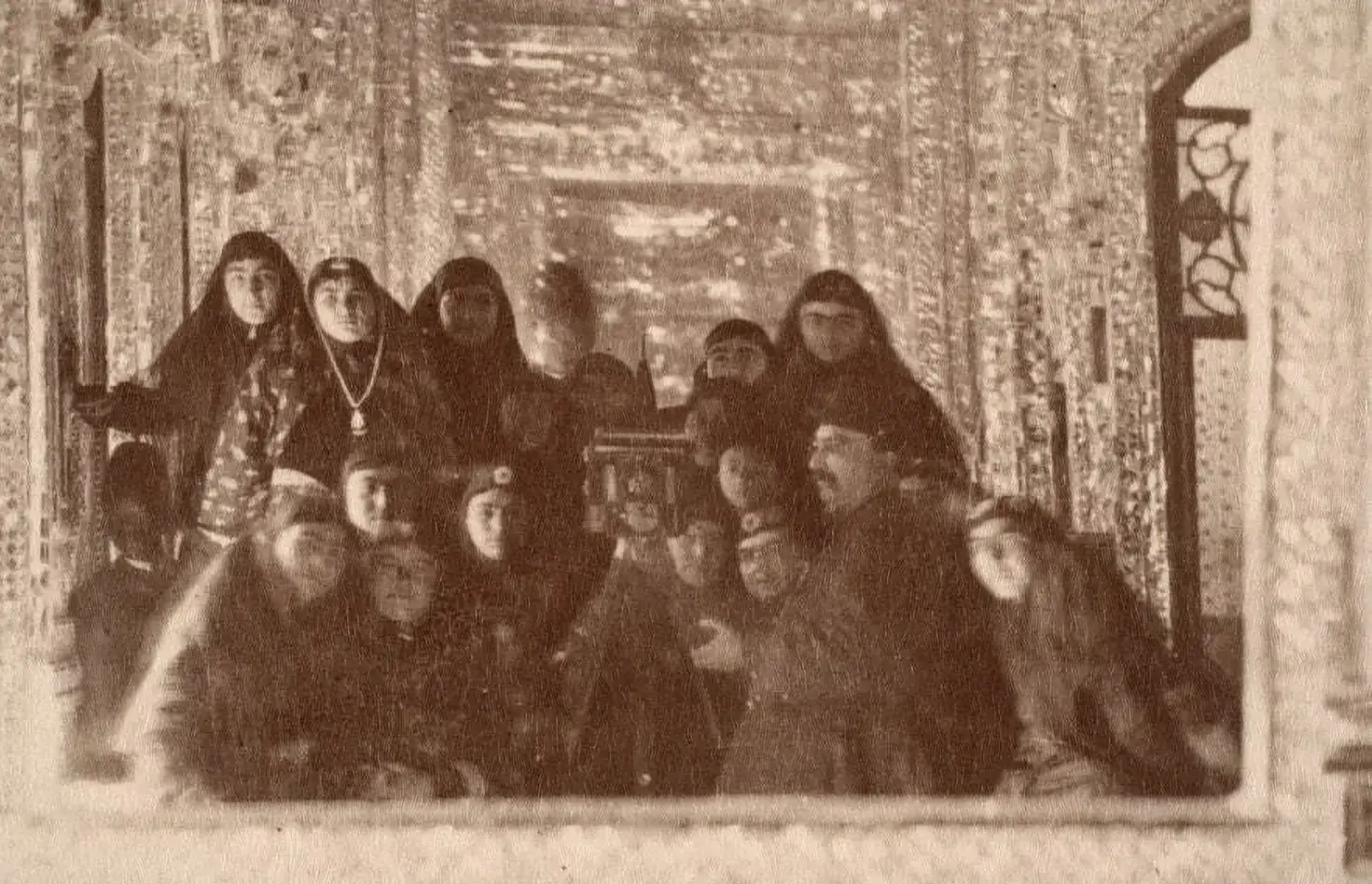 Persian emperor Naser al-Din Shah Qajar stands next to a camera, taking a picture into a mirror. He is standing surrounded by about 17 of his wives. They are in what appears to be a regal palace.