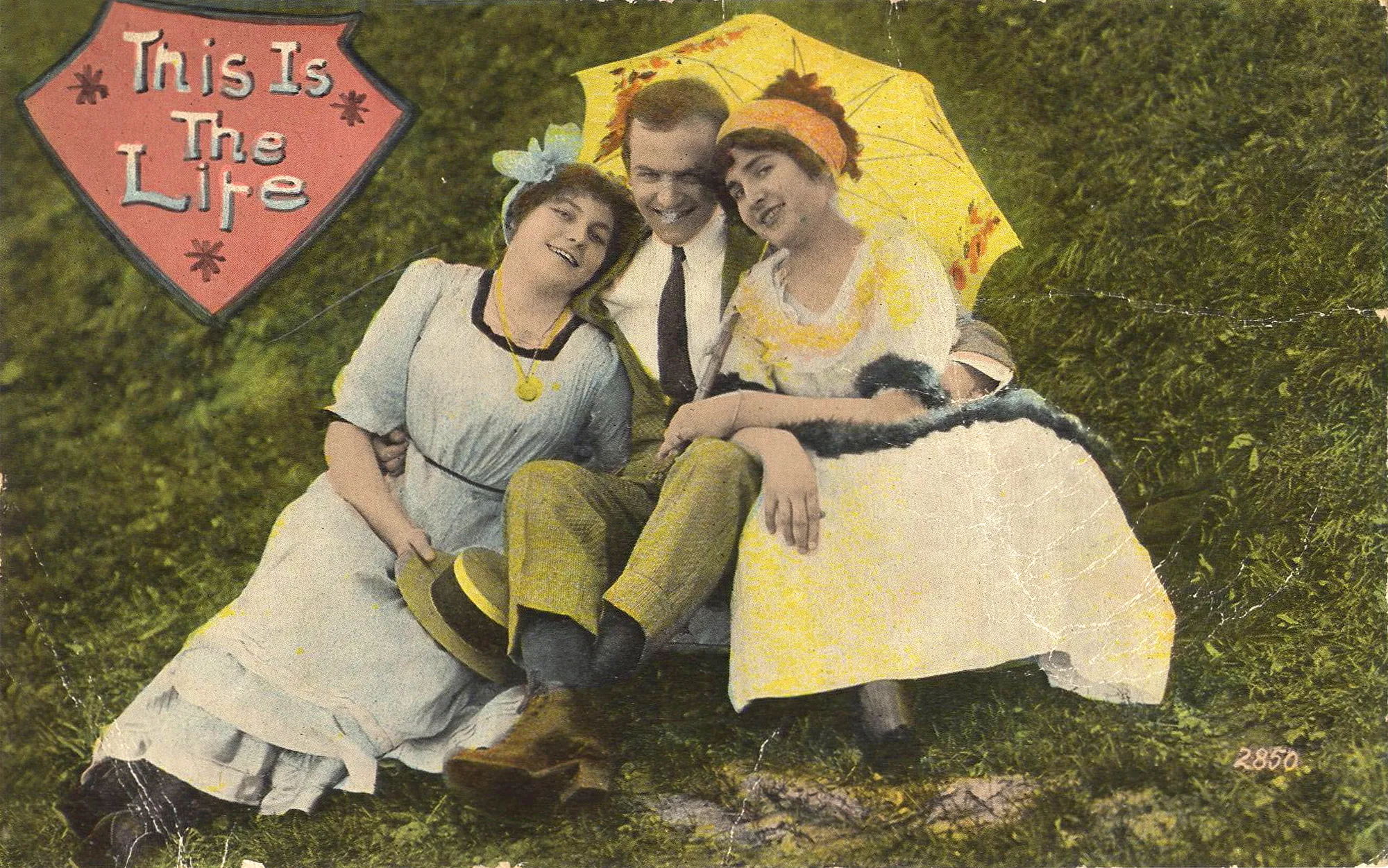 Postcard labeled “This Is The Life”, showing man between two women; the three are seated out-of-doors, all smiling, dressed in fashion of early 20th century. One woman holds a yellow parasol, the other the man’s straw boater hat.