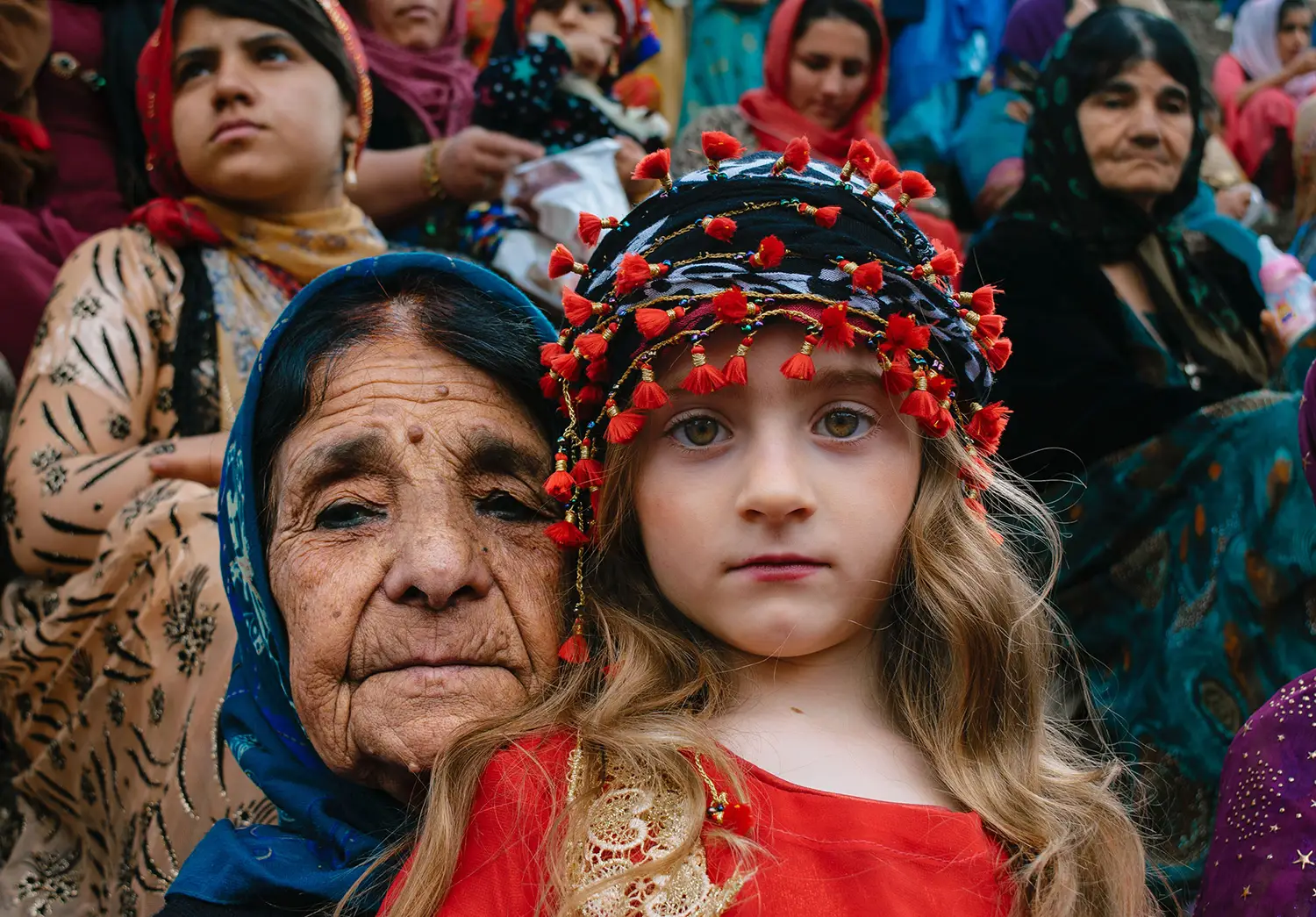 A grandmother and her grandchild watch the camera, as if deep in thought. The grandmother has deep set wrinkles, wears a blue headscarf, and leans close to the girl. The girl is wearing a red outfit with lace detailing. 