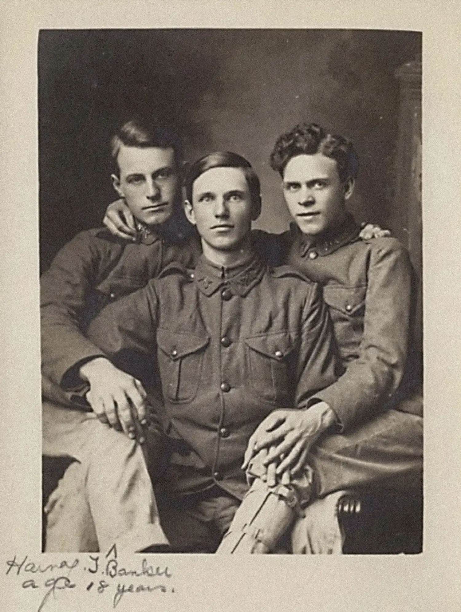 Three young men in WW1-era military uniform are seated together in an embrace. The man in the center is holding the hands of the two men seated on each side, and the outer two men each have a hand draped around the other’s shoulder.