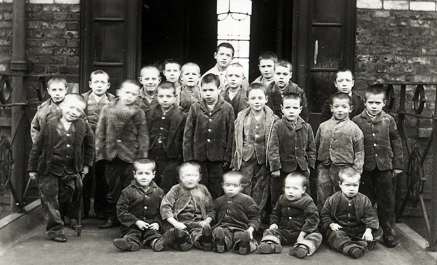 Twenty-three boys ranging from toddlers to about twelve years old are arranged for this group photograph. They all have their heads shaved and are wearing identical work clothes. The youngest sit in front, and one child appears to be missing a leg and using a cane. 