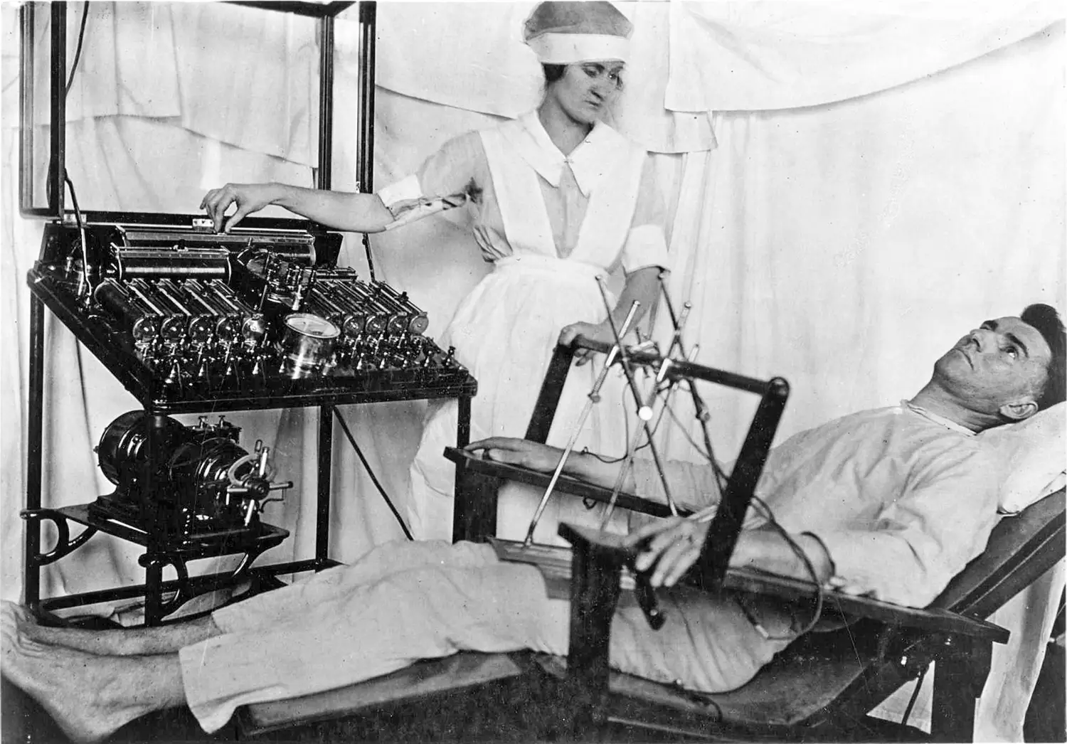 A nurse in a white uniform stands next to a complex mechanical apparatus with multiple tubes, dials, and gauges. She adjusts one of the settings with her hand. Lying on a reclined chair next to her is a male patient, attached to the machine via wires. 
