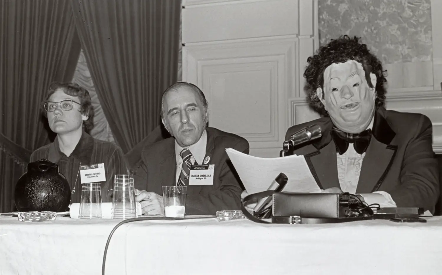 From left to right, a woman, man and person wearing a wig and mask are seated at a conference table. 