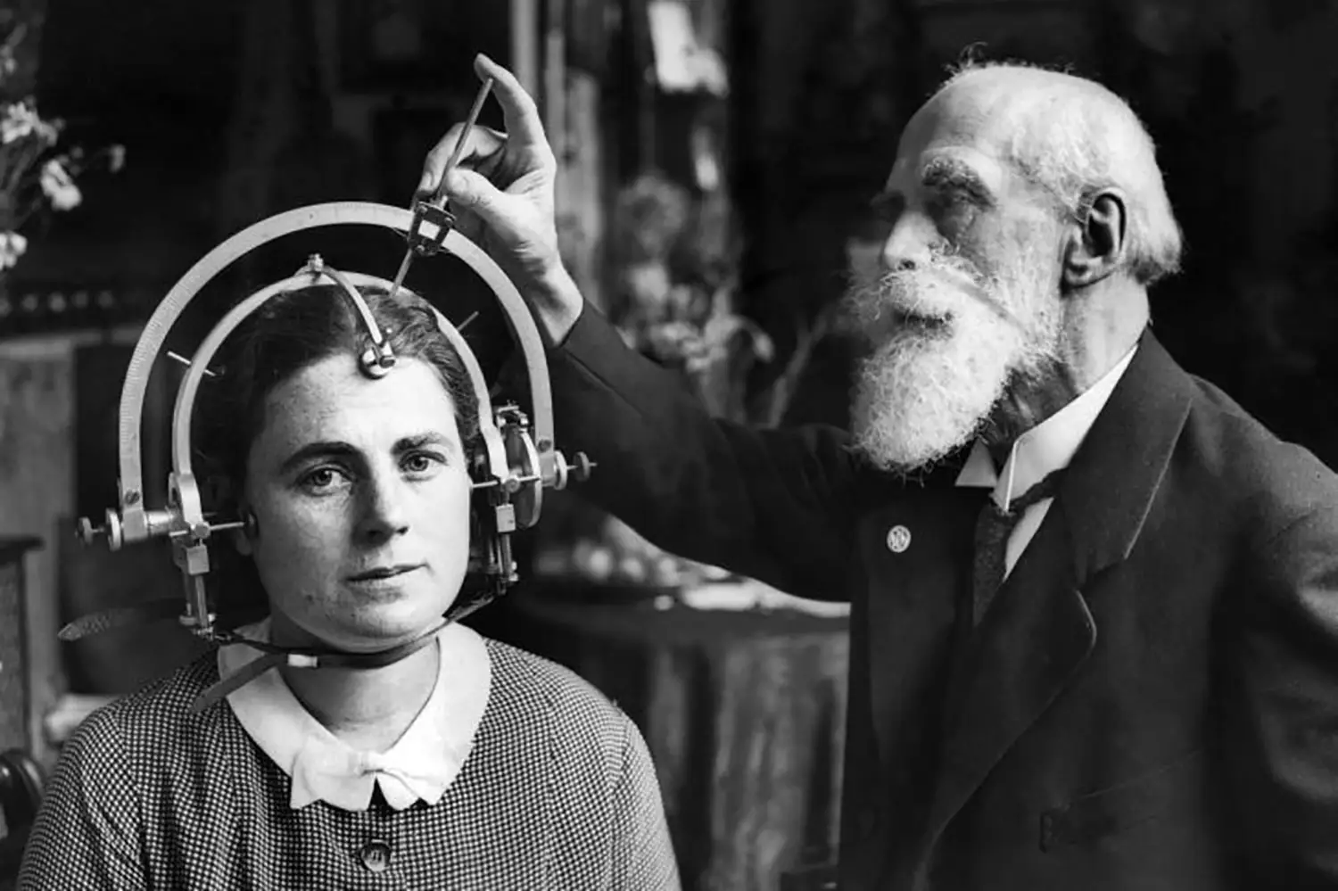 A woman sits with an elaborate circular measurement device strapped around her head. A much older bearded man adjusts the device while she stares into the camera.