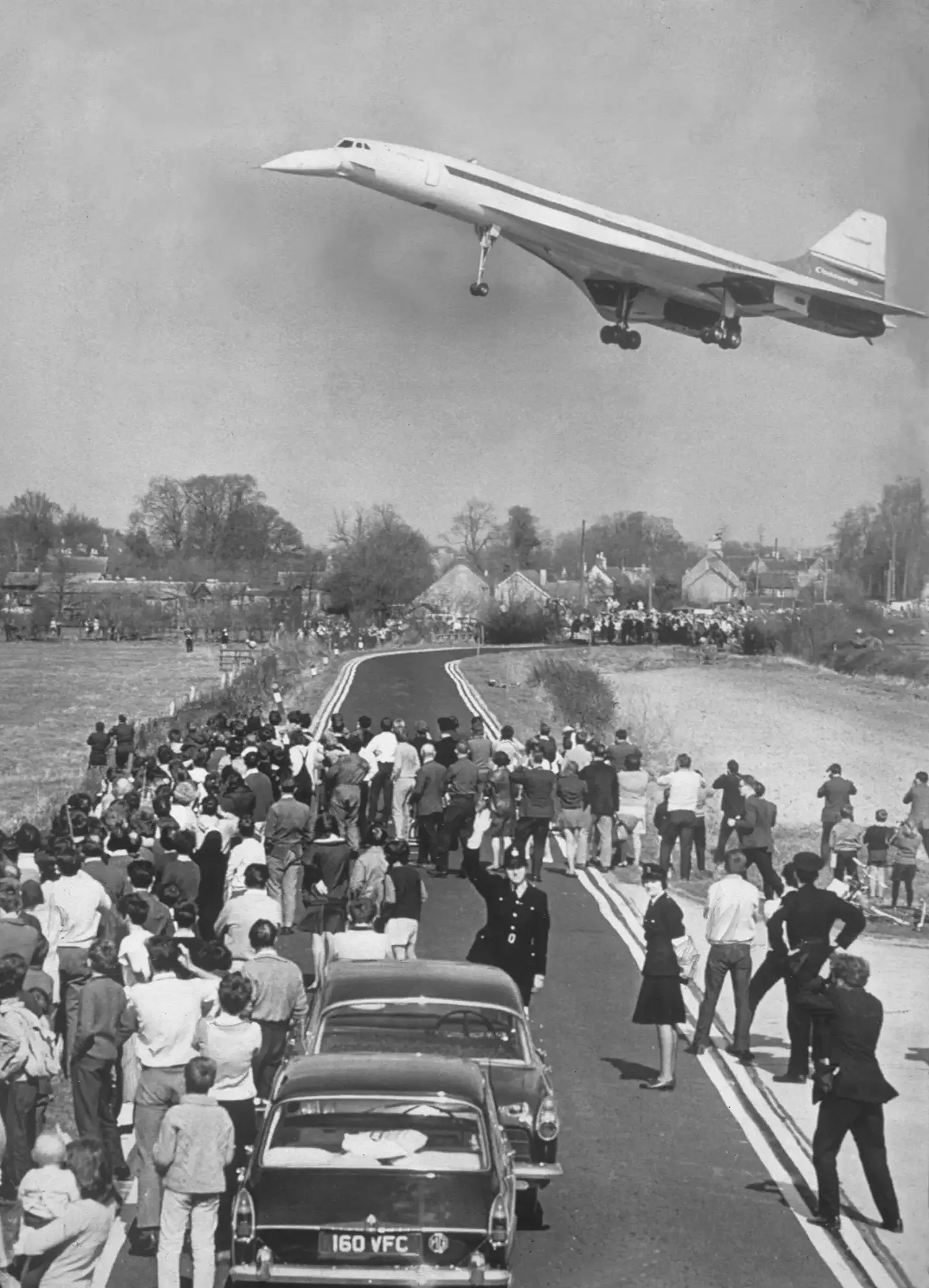 The Concorde aircraft flies low in the sky over a crowd of onlookers standing in the street below. The streamlined aircraft juxtaposes with the black and chrome mid-century cars stopped in the street.