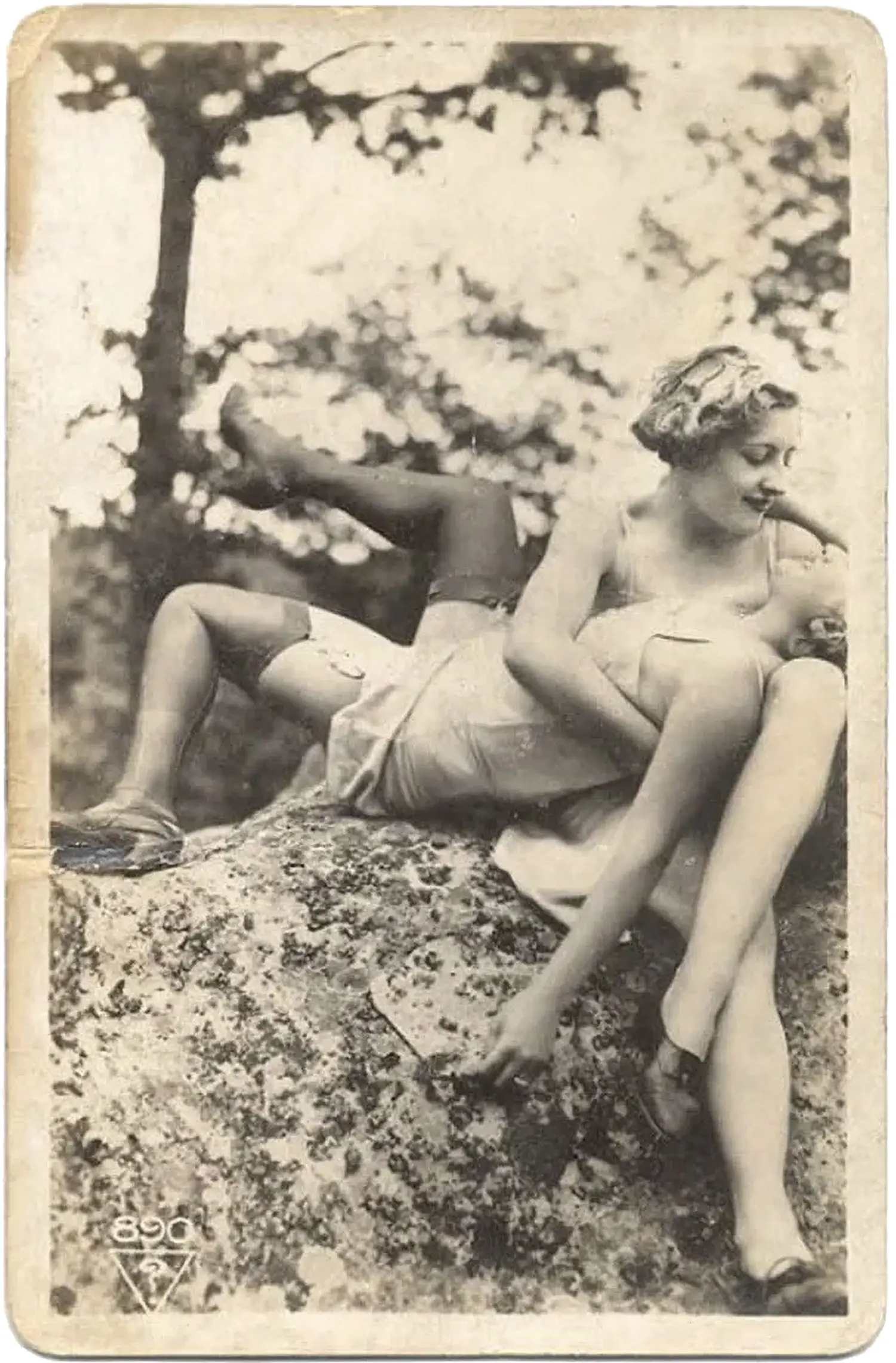 A weathered photograph of two women relaxing atop a large rock outdoors. One woman lies on her back, held by the other, who looks down at her face, smiling.