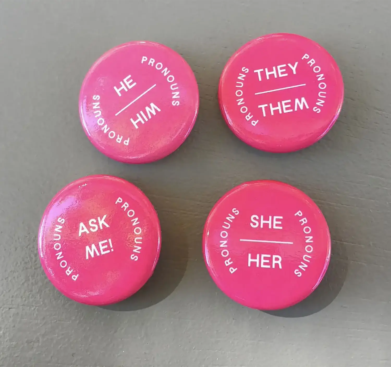 Four bright pink pronoun pins. Around the edge of each, it says “Pronouns,” in the center of each it says, respectively, “They/Them,” “She/Her,” “He/Him,” and “Ask Me!”