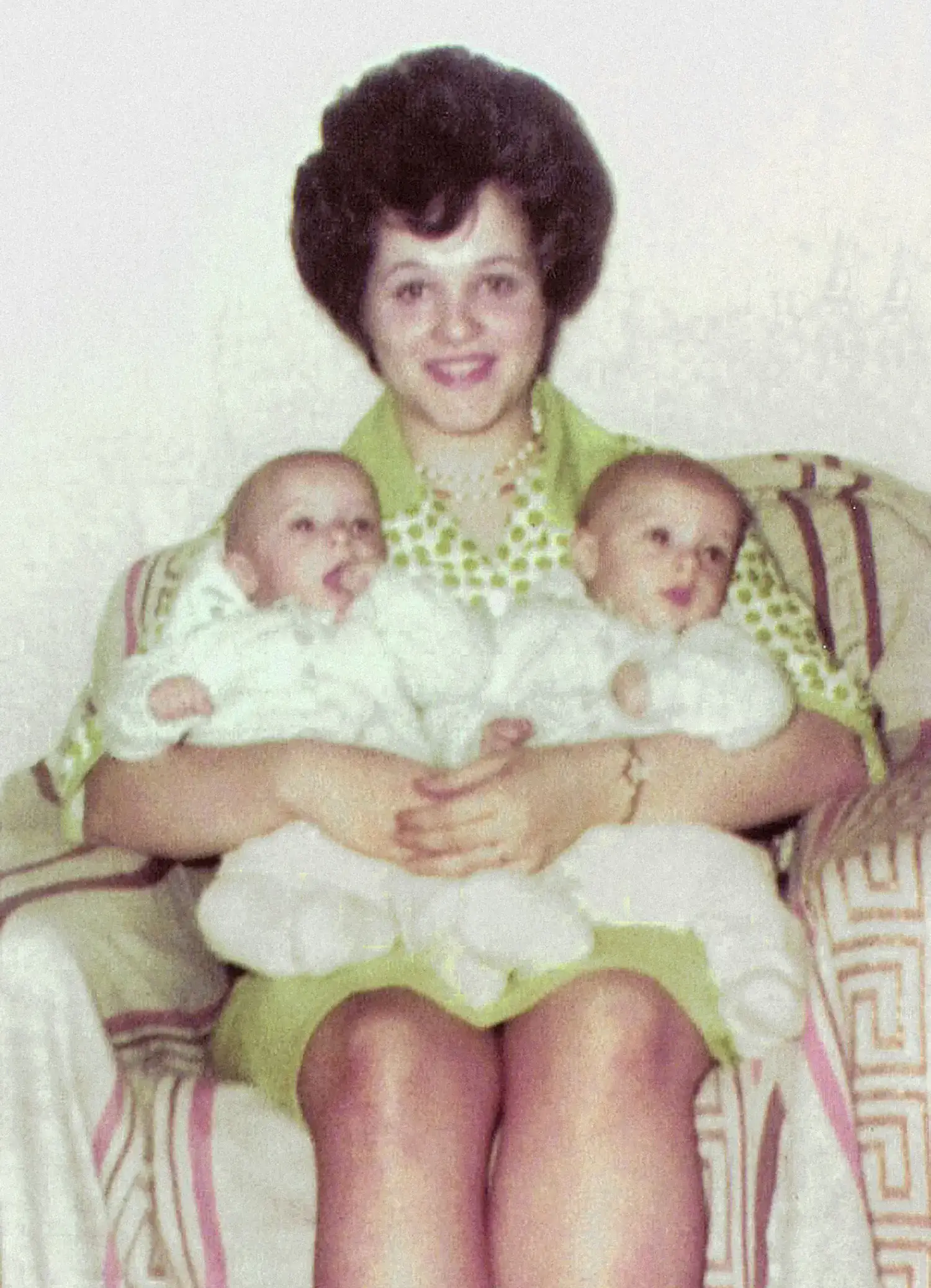 A woman, Janet Reimer, seated on a chair, smiling and holding two babies.