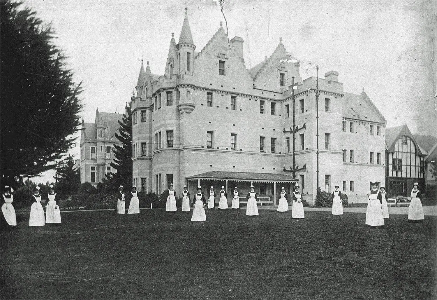 Nurses in white gowns stand spread on the lawn in front of an old castle-like structure that is the Seacliff Asylum.