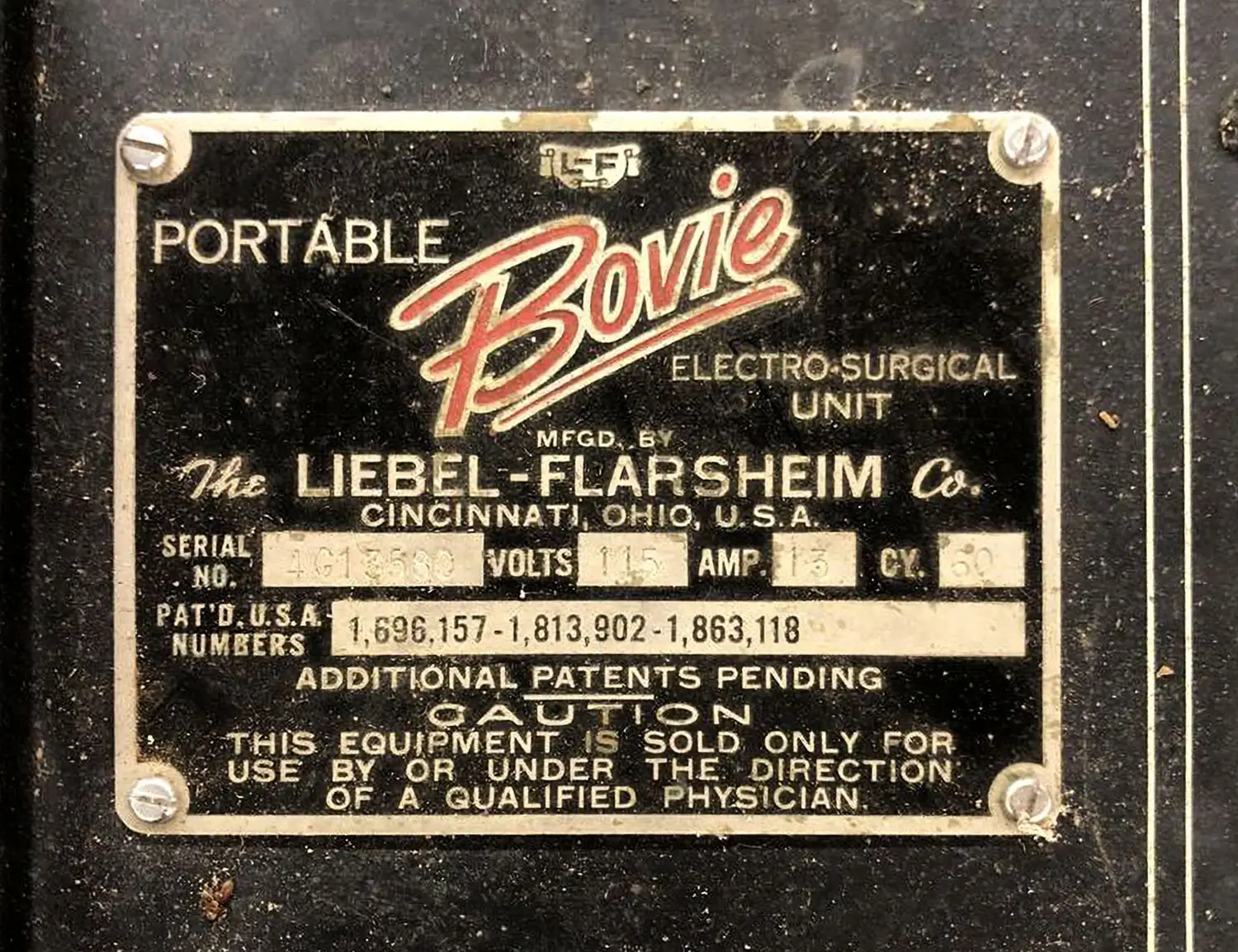 Black and gold metal nameplate that says “Portable Bovie Electro-Surgical Unit” and additional information related to its serial number, voltage, etc. It has a dirty and worn look.
