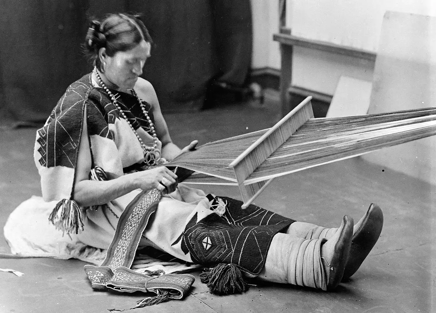 Wewa sits on the floor of the room, clad in traditional Zuni clothes and weaving a textile on a backstrap loom.