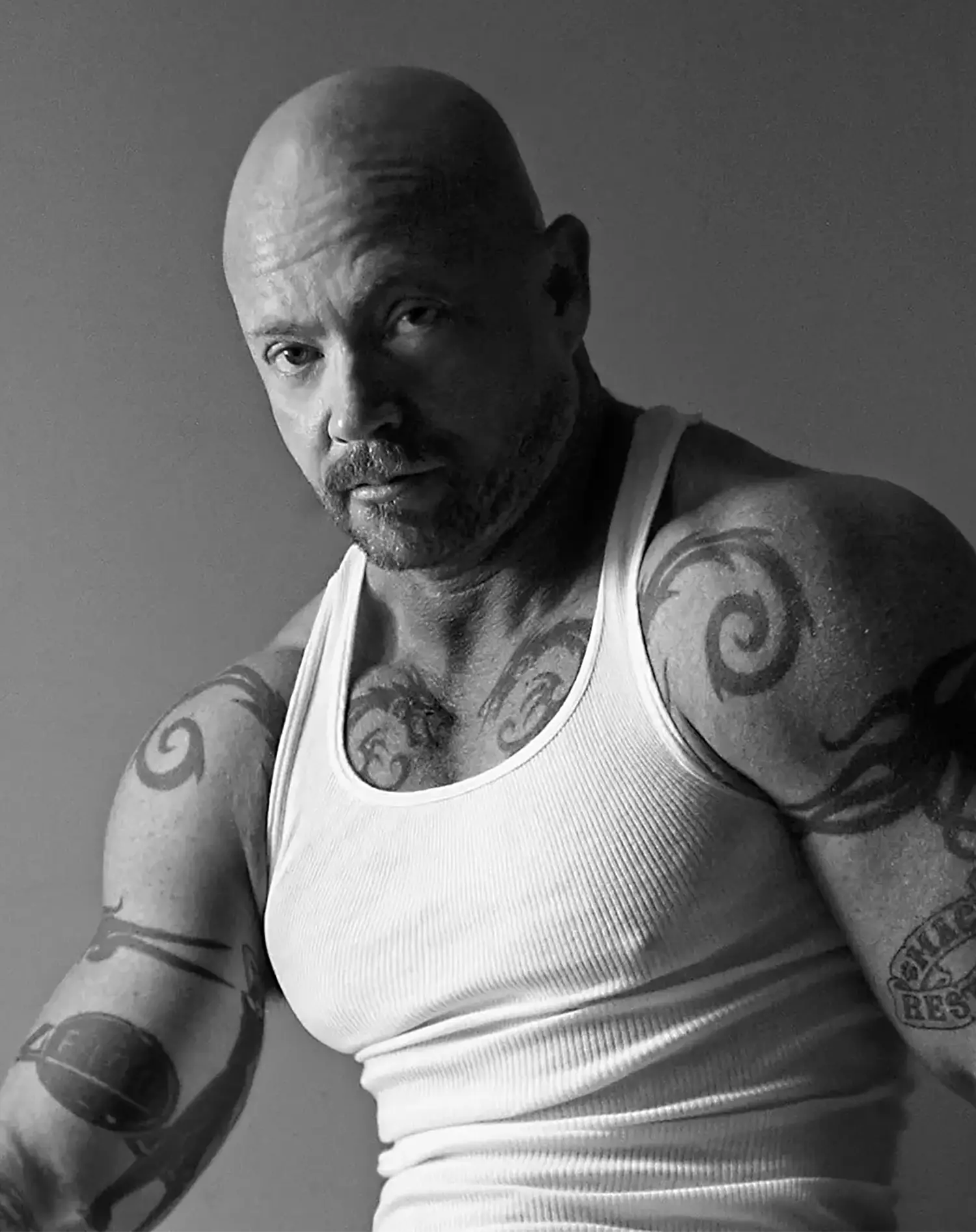 A studio portrait of a bald and bearded Buck Angel looking intently at the camera. He is wearing a white tank top that reveals arm and chest tattoos. 