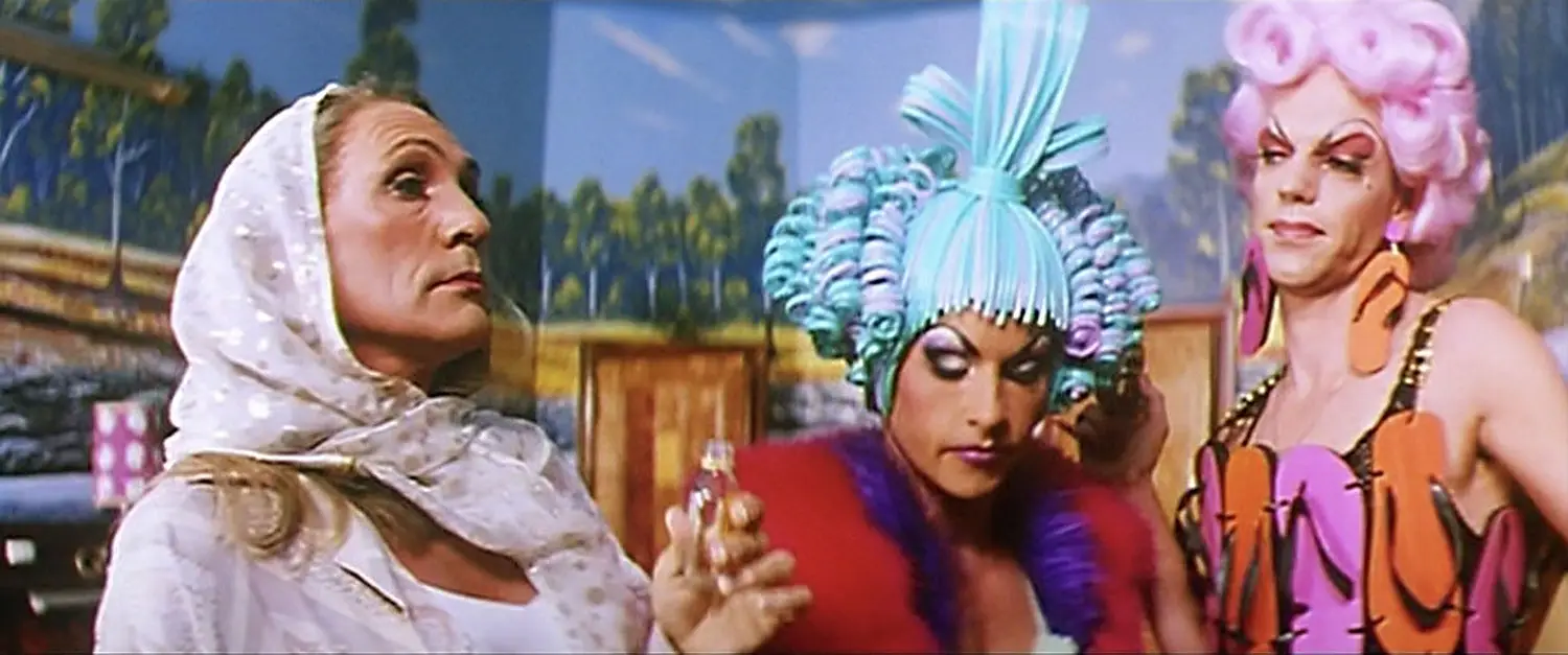 Movie still from The Adventures of Priscilla Queen of the Desert, showing the three main characters, one of which is trans and the other two who are dressed in colorful, flamboyant drag outfits.