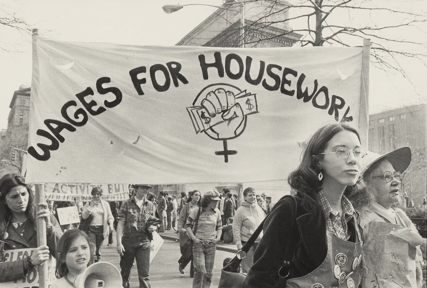 A group of protesters, primarily girls and women, marching with a banner that says “Wages for housework.”