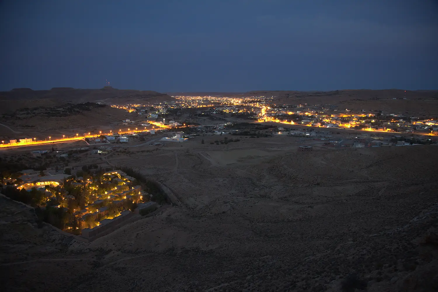 Dusk view of a barren desert landscape cut through with brightly lights indicating a settlement that carved downwards into the desert ground.