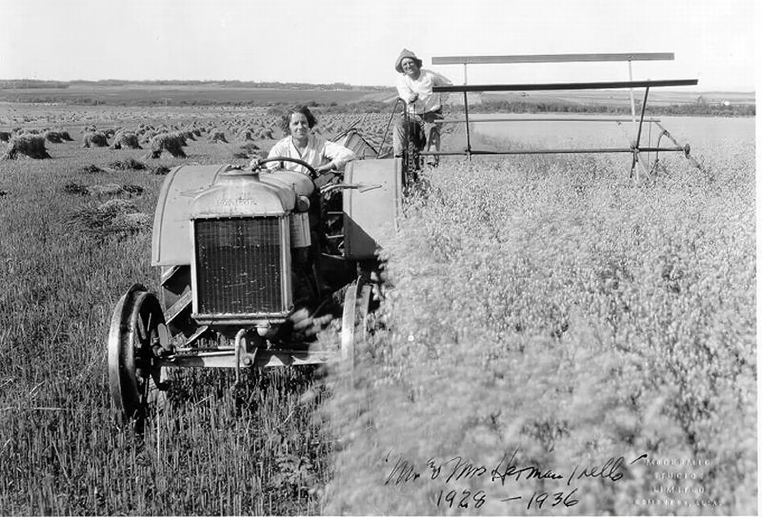 A woman drives a tractor. A man sits beside her, riding on an attached device that harvests the field.
