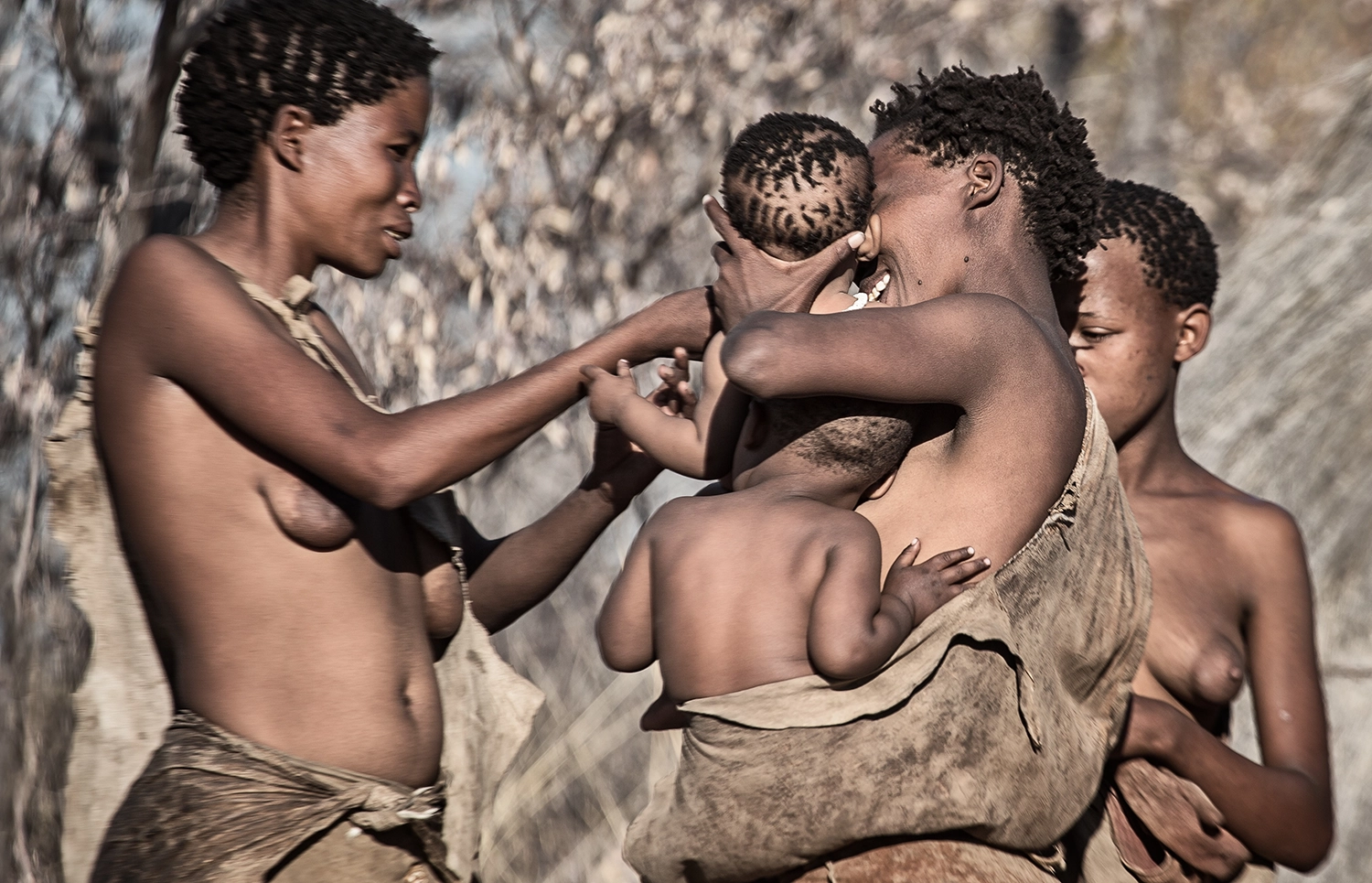 Mothers of the San people are gathered around a baby held by one of the women. That woman also carries another small child in a sling.
