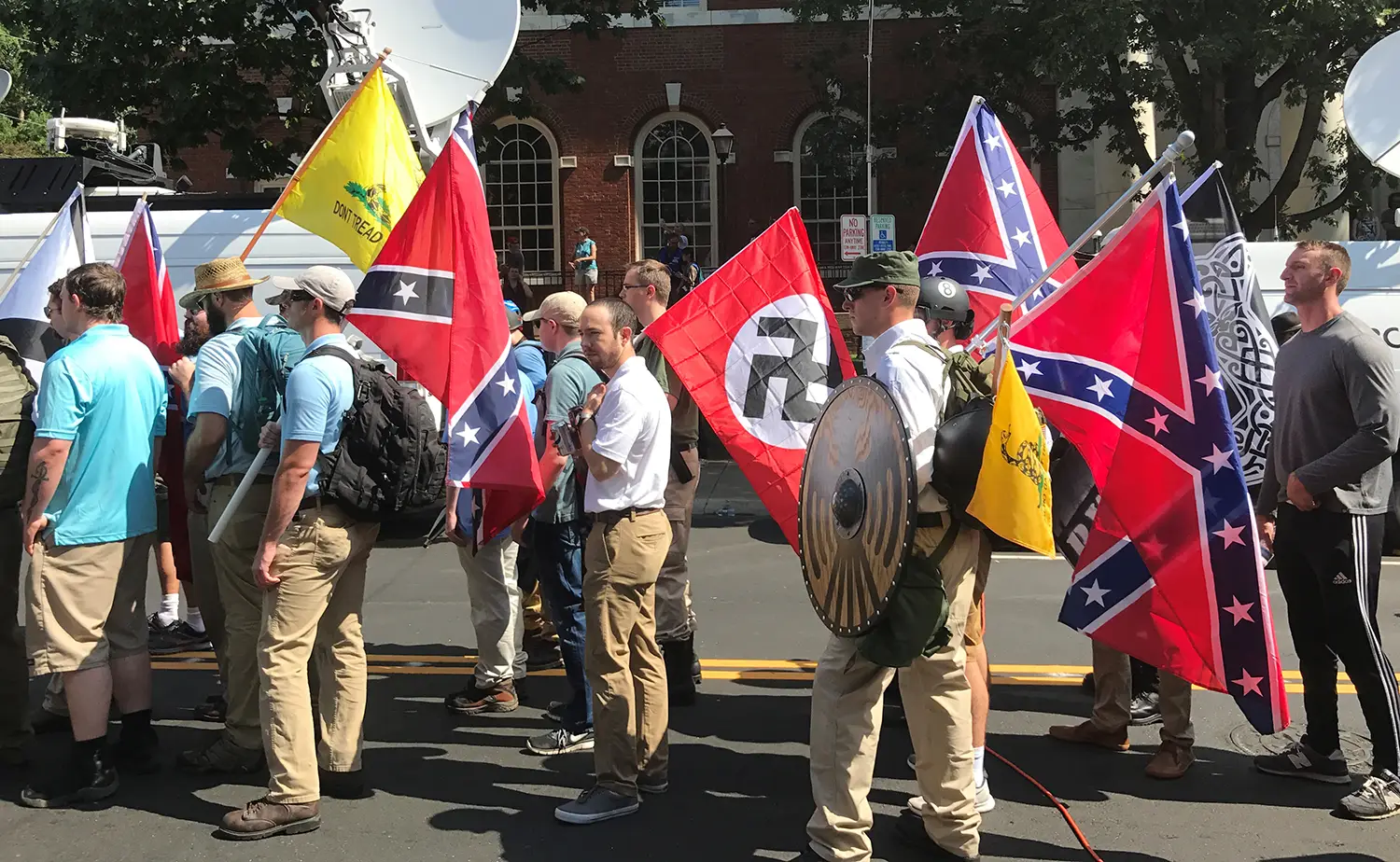 Various demonstrators, all male and wearing khaki pants, carrying flags with confederate and Nazi iconography. 