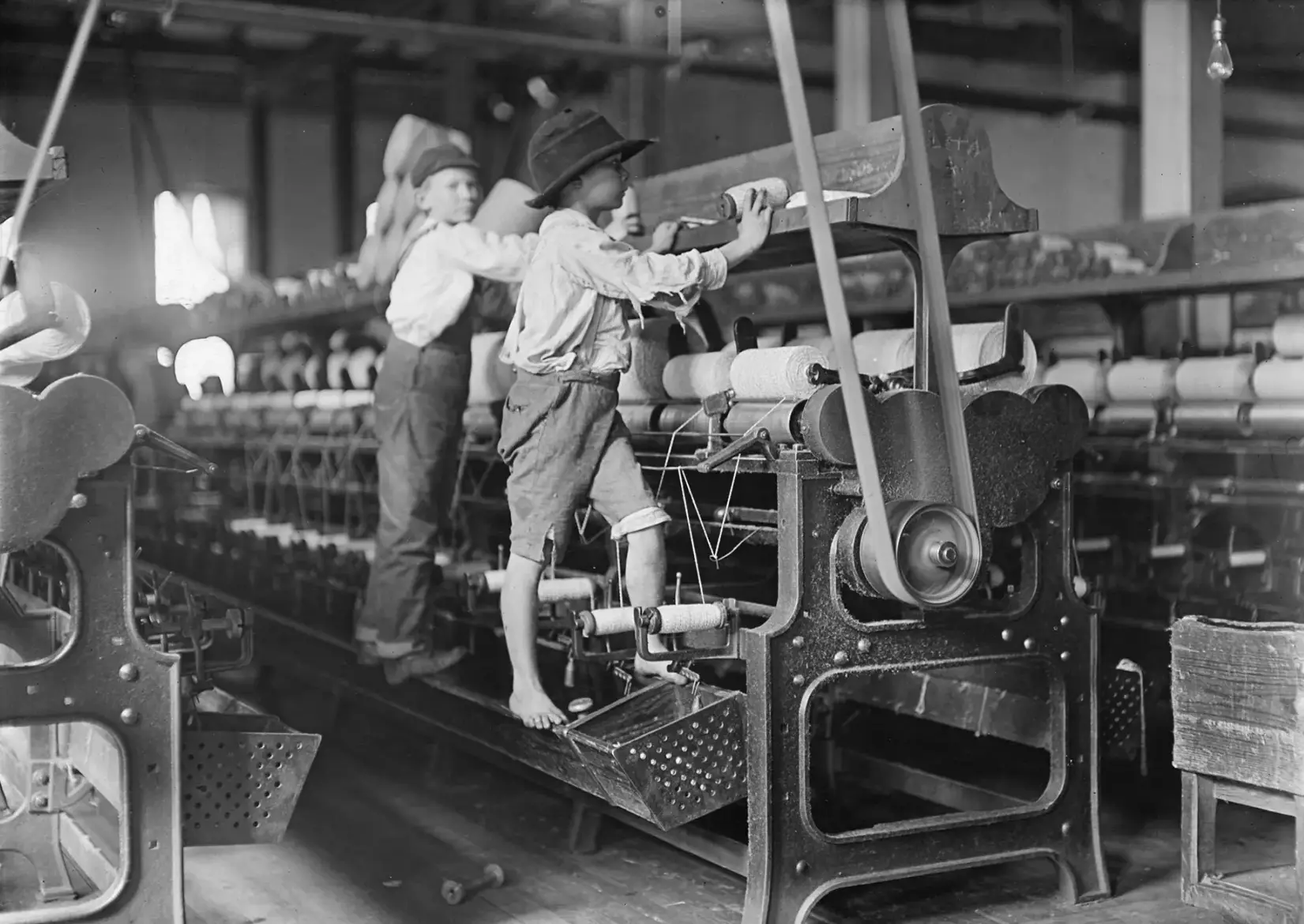 A large industrial textile machine, connected to a spinning belt, upon which stand two small, barefoot boys who are mending the machine.