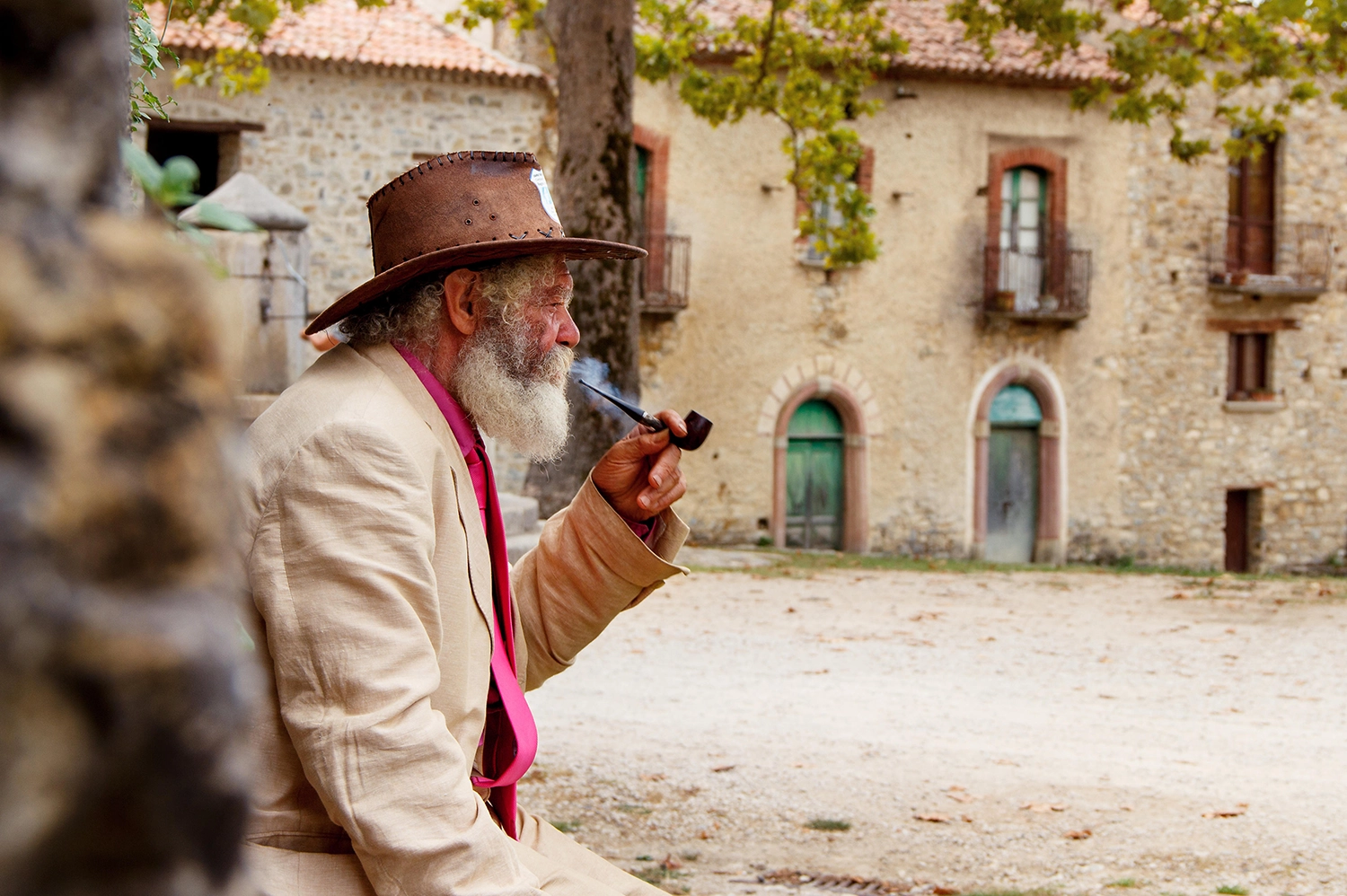 An old man sits alone in the plaza of an Italian village, smoking a pipe. 