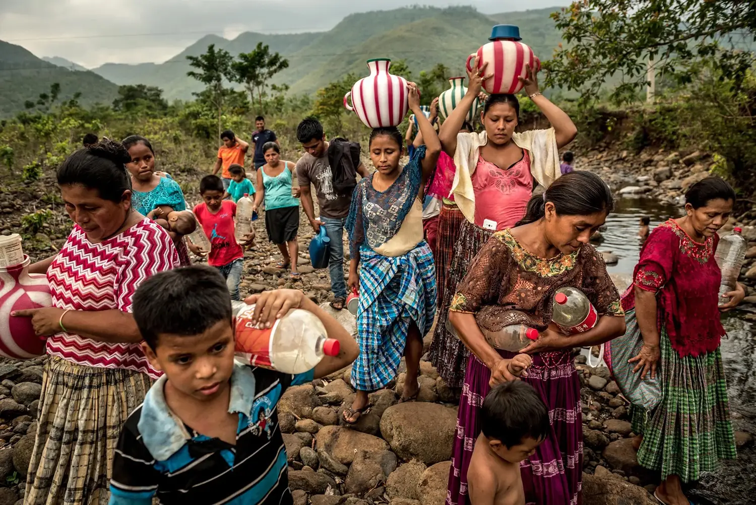 Women and children carry plastic bottles under arm and colorful jugs of water atop their heads. They are standing atop river rocks, and a small stream is visible in the background.