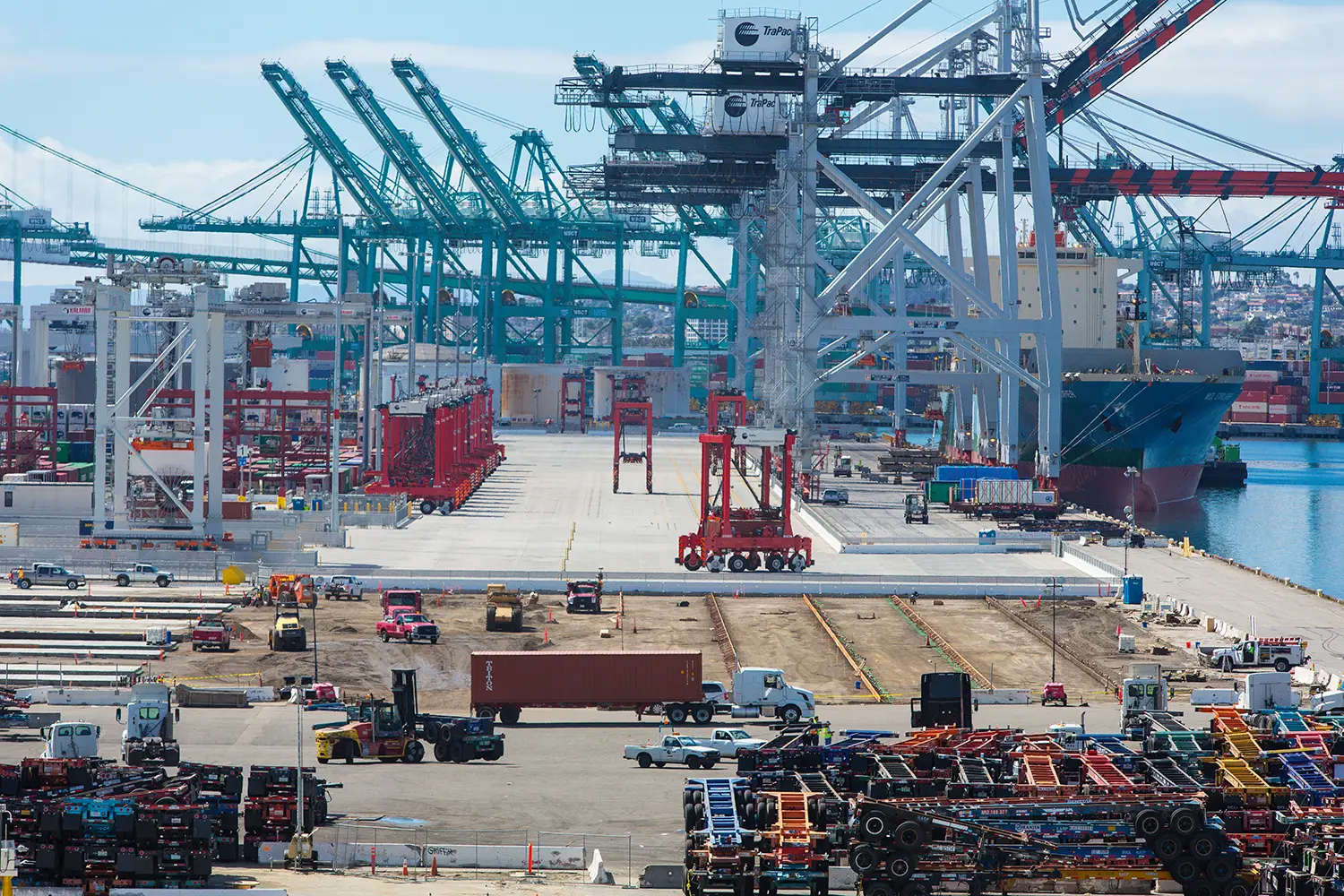 A bustling shipping port with large container cranes in the background. In front of them are tall red, automated straddle carriers.