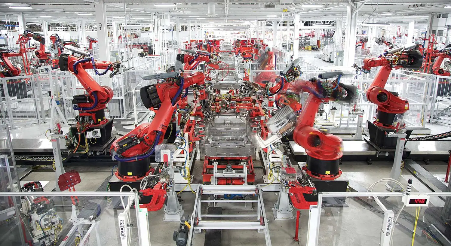 View of a factory floor. Large red robotic arms, in motion at work on vehicle chassis, are lined side by side until the back of the factory.
