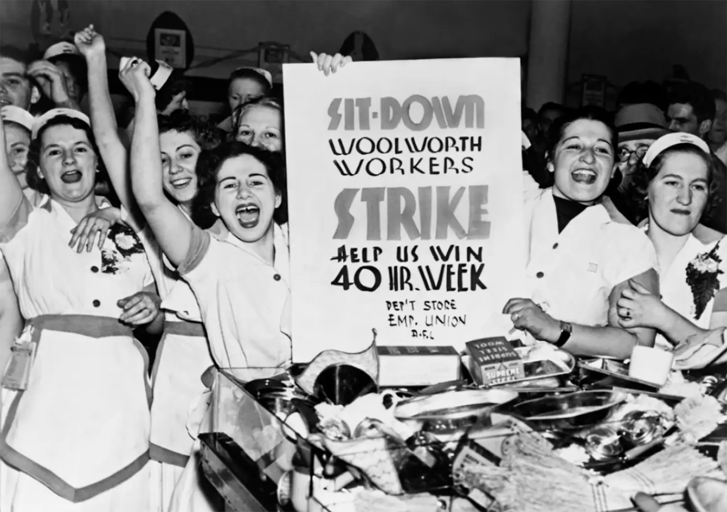 Women dressed in their Woolworth’s uniforms smile and cheer, holding the sign “Sit-down // Woolworth Workers Strike // Help us win 40 hr. week // Dep’t store emp. union.”